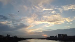 Sunset over a calm river with soft golden and blue clouds in the sky, a bridge in the distance, and city buildings silhouetted along the shoreline.

