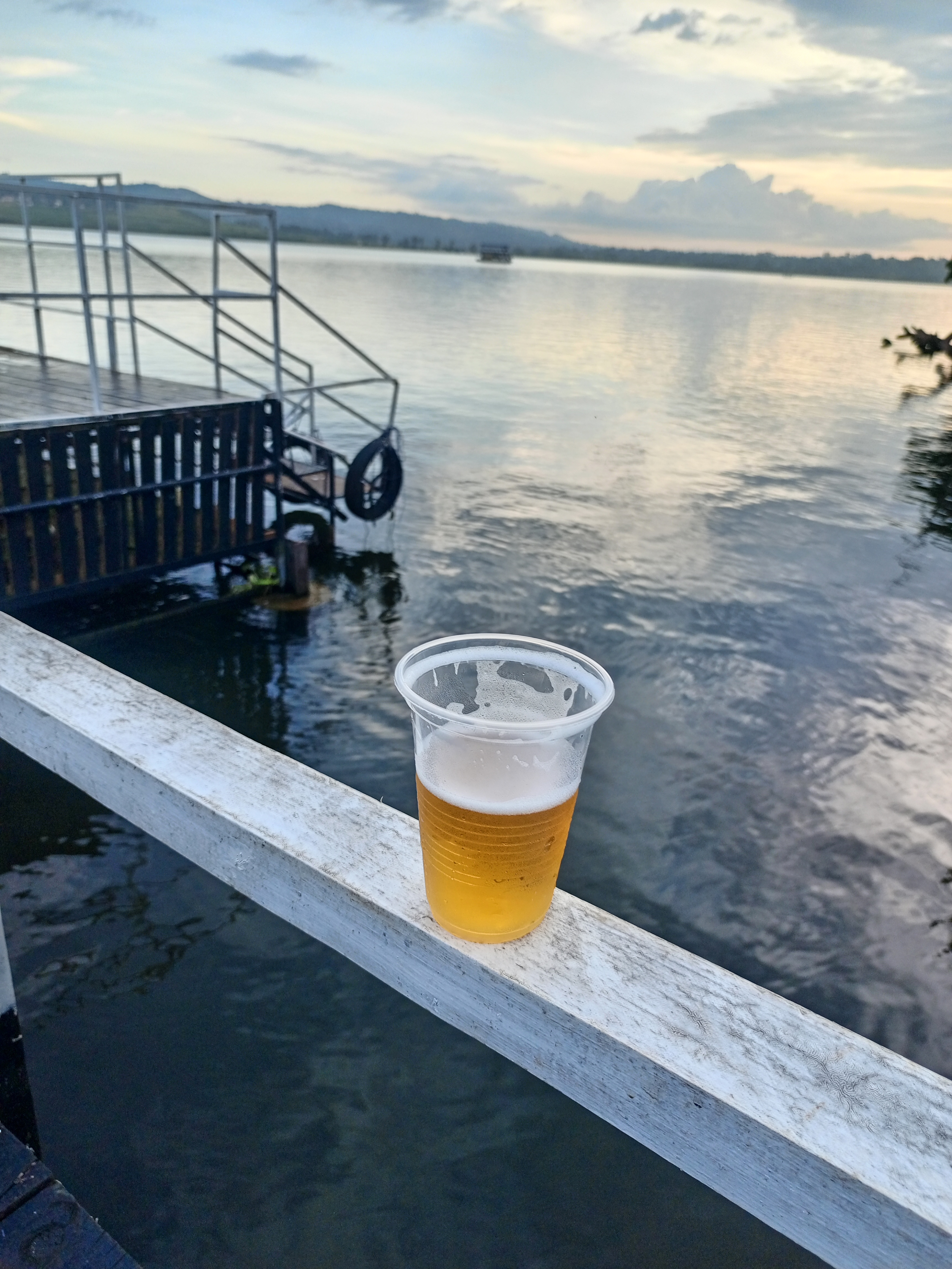 A clear plastic cup filled with golden beer sits on a white railing overlooking a calm body of water.