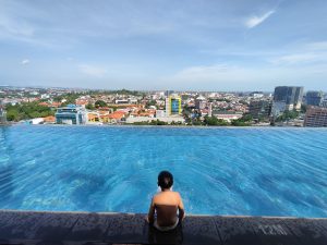 A person sitting at the edge of an infinity pool, overlooking a cityscape filled with a mix of modern buildings and traditional houses under a clear blue sky