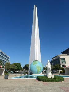 A tall white obelisk monument with a large globe sculpture and a human statue at its base under a clear blue sky