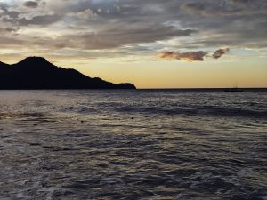 A tranquil seascape at sunset, featuring calm waters reflecting the golden and orange hues of the sky. In the distance, a small fishing boat is gently sailing near the horizon, while a dark silhouette of a hill stands on the left side of the image against a backdrop of scattered clouds