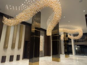 A modern, spacious interior with high ceilings and hanging flower-like decorations in orange and white. Soft lighting creates a warm feel, with textured walls, gold-accented pillars, glass panels, and polished white marble flooring.
