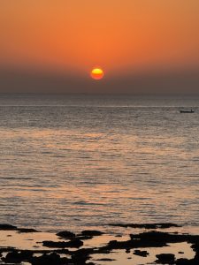 Vertical shot of a deep orange sunset over the sea. A glowing yellow-orange sun is centered above the horizon line. In the foreground, dark silhouettes of rocks are visible in the shallow water, reflecting the warm colors of the sky. A tiny silhouette of a boat sits on the right side of the horizon.