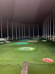 A brightly illuminated golf driving range at night, featuring multiple colorful target areas on the green turf. Tall poles rise in the background against a cloudy sky, and the scene is lit by artificial lights, creating a vibrant atmosphere. A protective netting can be seen in the foreground.