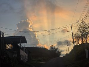 A scenic view of a road leading uphill, framed by a house on the left and power lines overhead. In the background, a dramatic sky features fluffy clouds illuminated by the warm hues of sunset, with rays of light streaming through them. The setting conveys a tranquil, picturesque atmosphere.