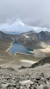 From the crater rim of Nevado de Toluca, the twin lakes — Laguna del Sol and Laguna de la Luna — fill the basin below, with cinder cones dividing the blue water and the Toluca Valley stretching to the horizon.
