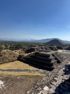 A panoramic view of ancient pyramids at Teotihuacan, showcasing stepped structures set against a clear blue sky. In the foreground, a series of smaller pyramids are visible, while a larger pyramid rises prominently in the background. The surrounding landscape features sparse vegetation and distant mountains, creating a serene atmosphere.
