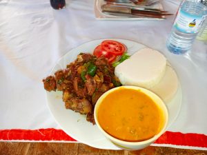 A plate of sautéed beef with peppers and onions is served with slices of ugali, a bowl of peanut sauce, and fresh tomato and lettuce.