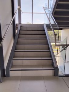 A modern staircase featuring light gray tiles, leading up with smooth edges and a stainless steel handrail. 