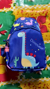 A small blue kids’ backpack with a colorful dinosaur print, placed on a floral tablecloth.