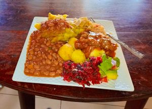 Generous portions of stewed brown beans and mashed yellow matooke cover half of a white textured plate. The meal includes beef stew, boiled potatoes, a side of sautéed buuga (red amaranthus), and fresh avocado slices. A stainless steel fork rests on the side of the plate, which is positioned on a dark, polished wooden dining table.