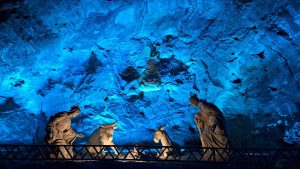 A nativity scene carved inside the Zipaquirá Salt Cathedral, with white figures of Mary, Joseph, an angel, and animals glowing against a dramatic blue-lit salt rock wall, creating a sacred underground atmosphere.