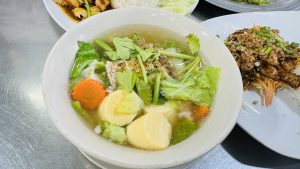 A white bowl of clear vegetable soup with tofu, carrots, and greens, garnished with cilantro, served alongside Thai dishes on a metal table under bright indoor lighting.
