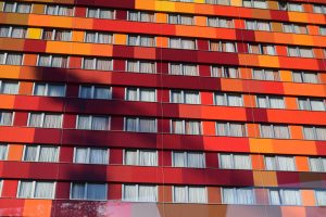 A close-up view of a modern building facade featuring a grid pattern with vibrant multi-colored panels in red, orange, and yellow. Includes multiple rows of uniform windows.