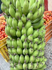 A close-up of green bananas stacked tightly, with red tomatoes in crates behind them in a market setting.