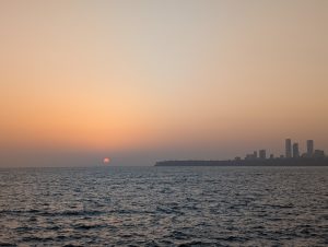 A serene sunset over the ocean, with the sun partially dipped below the horizon, casting a warm glow in shades of orange and pink. The calm waters reflect the colors of the sky, while a city skyline with tall buildings is visible in the distance along the right side of the image. The overall atmosphere is peaceful and picturesque, evoking a sense of tranquility.
