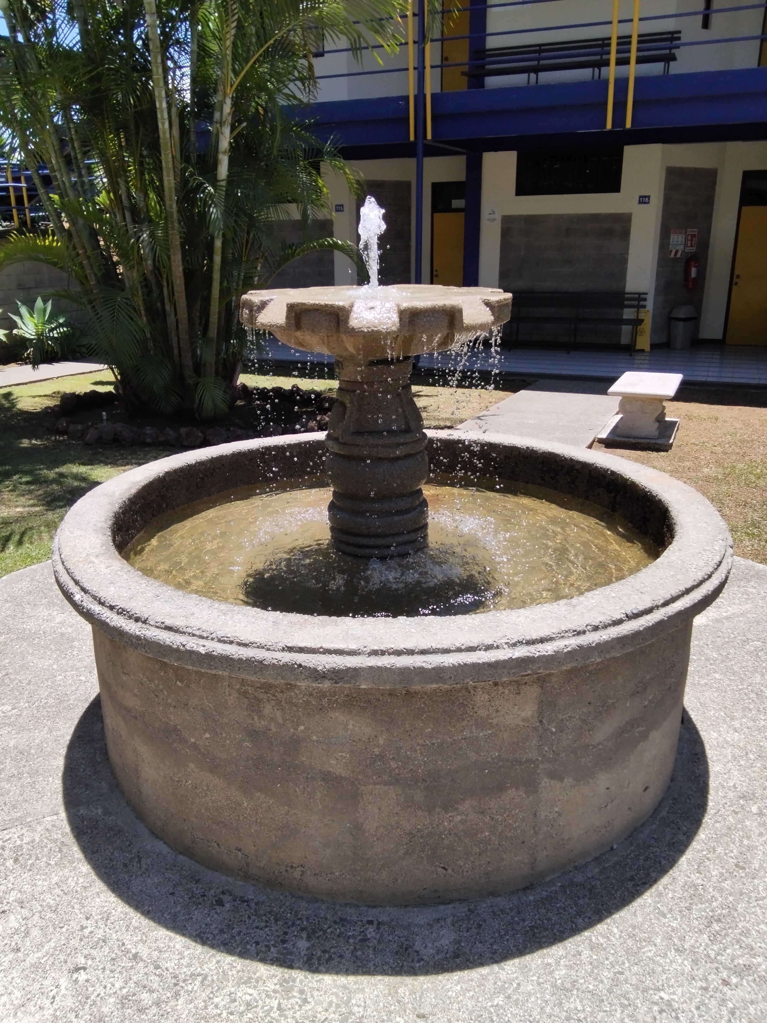 A circular stone fountain with a tiered design is the central feature, with water spraying upward from the top tier.