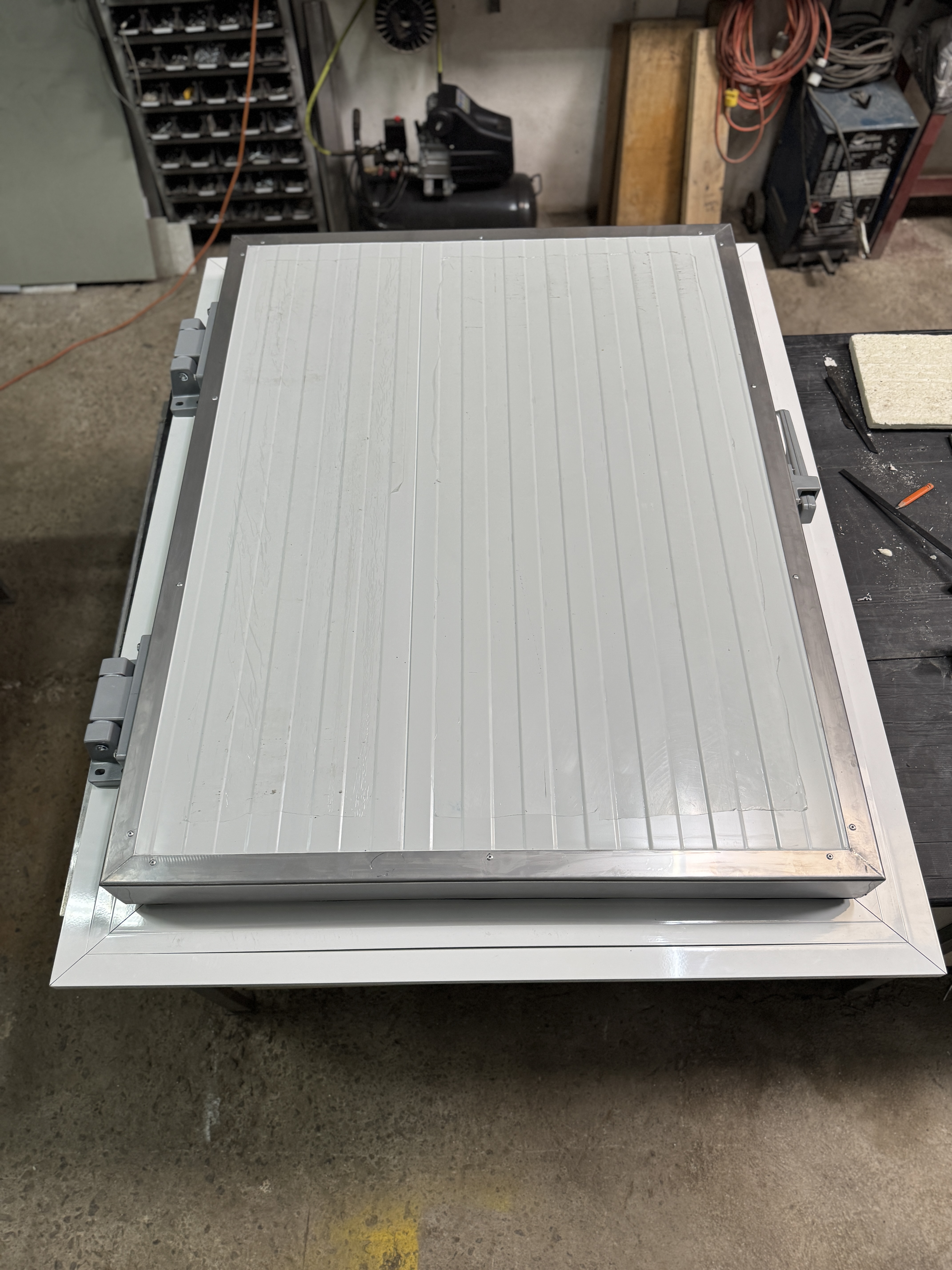 A flat, rectangular surface with a ribbed texture is shown, featuring a metal edge and hinges on one side. The surface is light gray, and it is placed on top of a white table. In the background, there is a shelving unit filled with small compartments and some industrial equipment, including a compressor and tools. The floor appears to be concrete, suggesting a workshop or industrial setting.