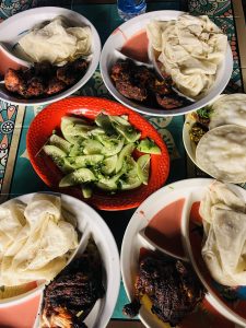 Four plates of grilled chicken and flatbreads surround a red dish of sliced cucumbers and lime. The setting has a vibrant, casual feel.