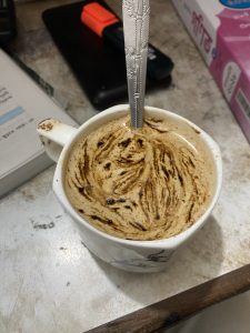 A creamy cappuccino with swirls of frothy foam in a white mug, a spoon inside, on a cluttered desk with a book, highlighter, and papers nearby.
