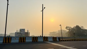 Wide shot of a paved road and bridge at hazy sunrise, with street lamps, striped barriers, an unfinished building, and distant trees under a pale golden sky.