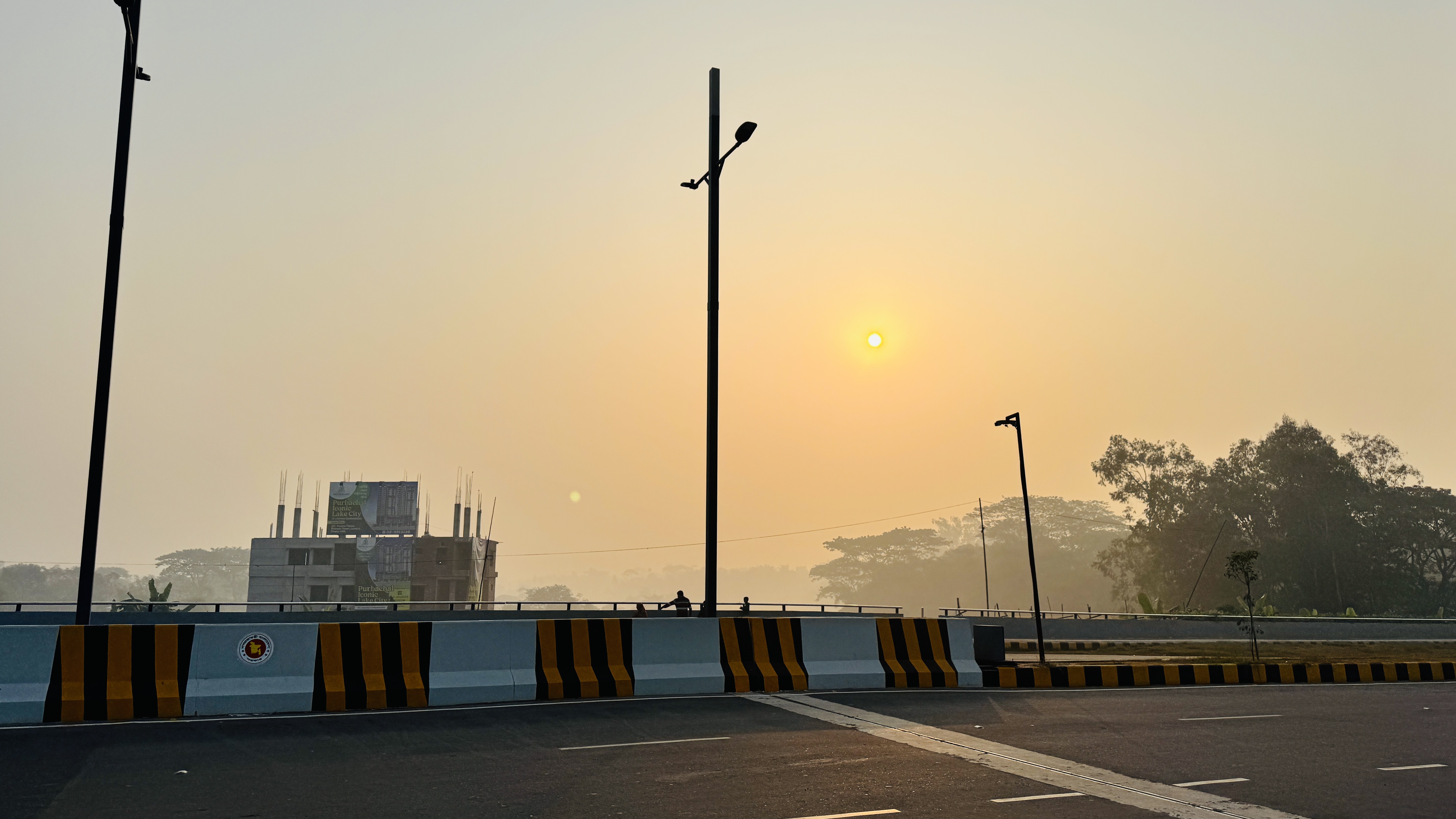 Wide shot of a paved road and bridge at hazy sunrise, with street lamps, striped barriers, an unfinished building, and distant trees under a pale golden sky.