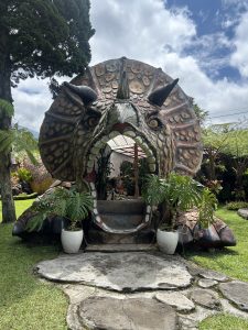 A large, detailed sculpture of a horned dinosaur head with an open mouth forming an entrance. The pathway leads inside through its jaws, surrounded by lush greenery, potted plants, and trees under a partly cloudy sky.