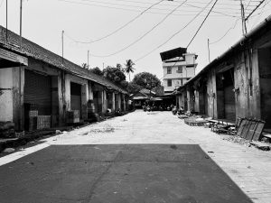 A black and white photo of a deserted market street featuring several closed shop fronts with rolling metal shutters, crates stacked outside, and scattered debris on the ground. Calicut old vegetable market.