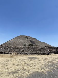 A large pyramid structure rises against a clear blue sky, partially shaded by a rocky wall in the foreground. The pyramid features distinct staircases leading up its side, with sparse vegetation growing on its surface. The ground is dry and mostly barren with patches of brown and yellow grass. A small figure can be seen sitting near the wall, adding scale to the ancient monument.