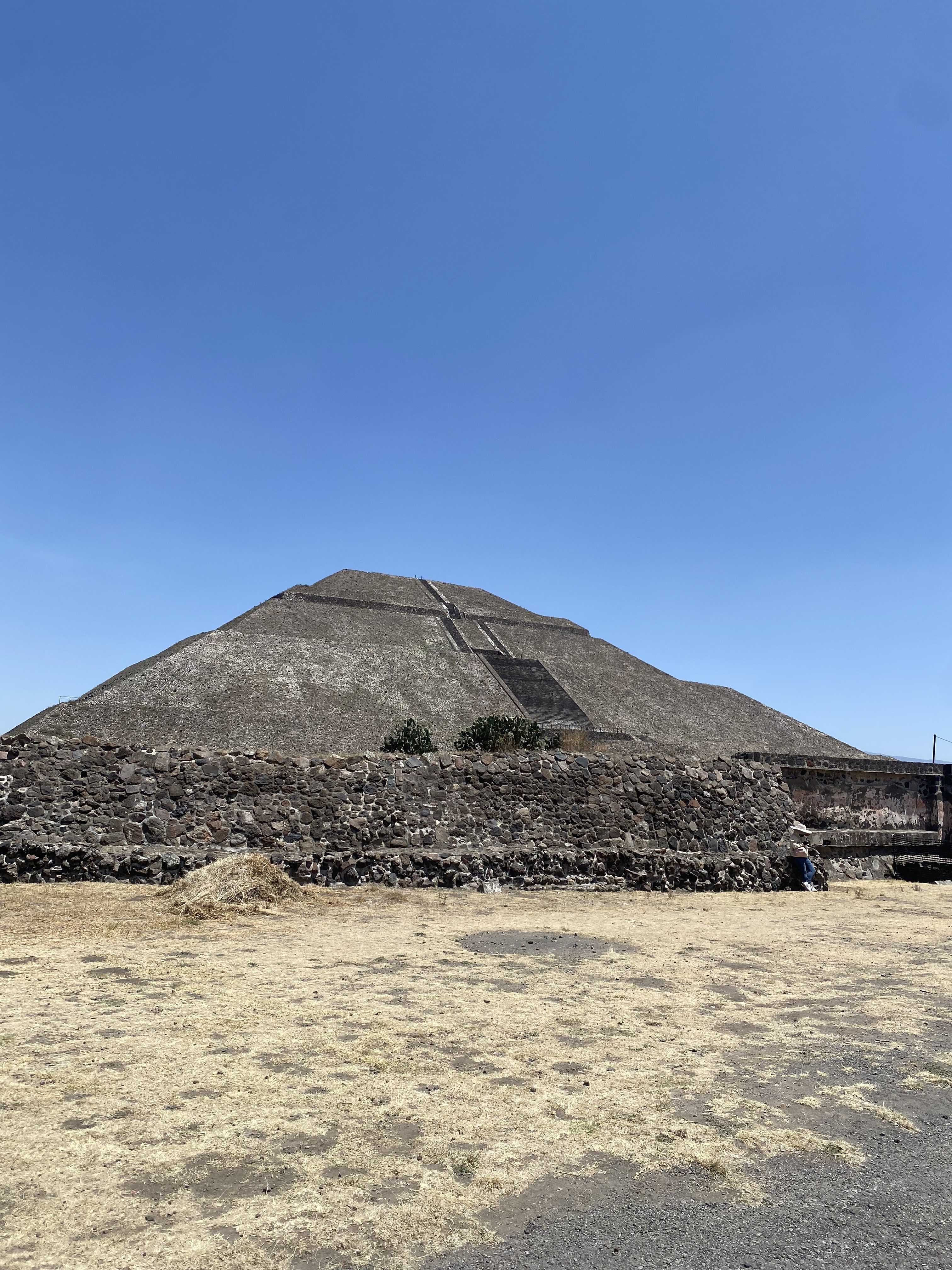 A large pyramid structure rises against a clear blue sky, partially shaded by a rocky wall in the foreground. The pyramid features distinct staircases leading up its side, with sparse vegetation growing on its surface. The ground is dry and mostly barren with patches of brown and yellow grass. A small figure can be seen sitting near the wall, adding scale to the ancient monument.