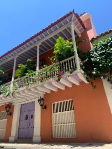 A beautiful colonial building painted in salmon pink with a white balcony overflowing with tropical plants and flowers