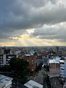 Crepuscular rays break through a dramatic storm sky over Bogotá, Colombia, bathing the sprawling city in warm golden light. A dense mix of brick residential buildings, mid-rise offices and mature trees stretches westward toward the flat savanna in the distance, with a TransMilenio bus visible on the street below.