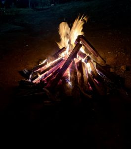 A brightly burning campfire made of stacked wooden logs, with flames flickering upwards against a dark background. The fire emits a warm glow, illuminating the surrounding area slightly, while shadows are cast on the ground.