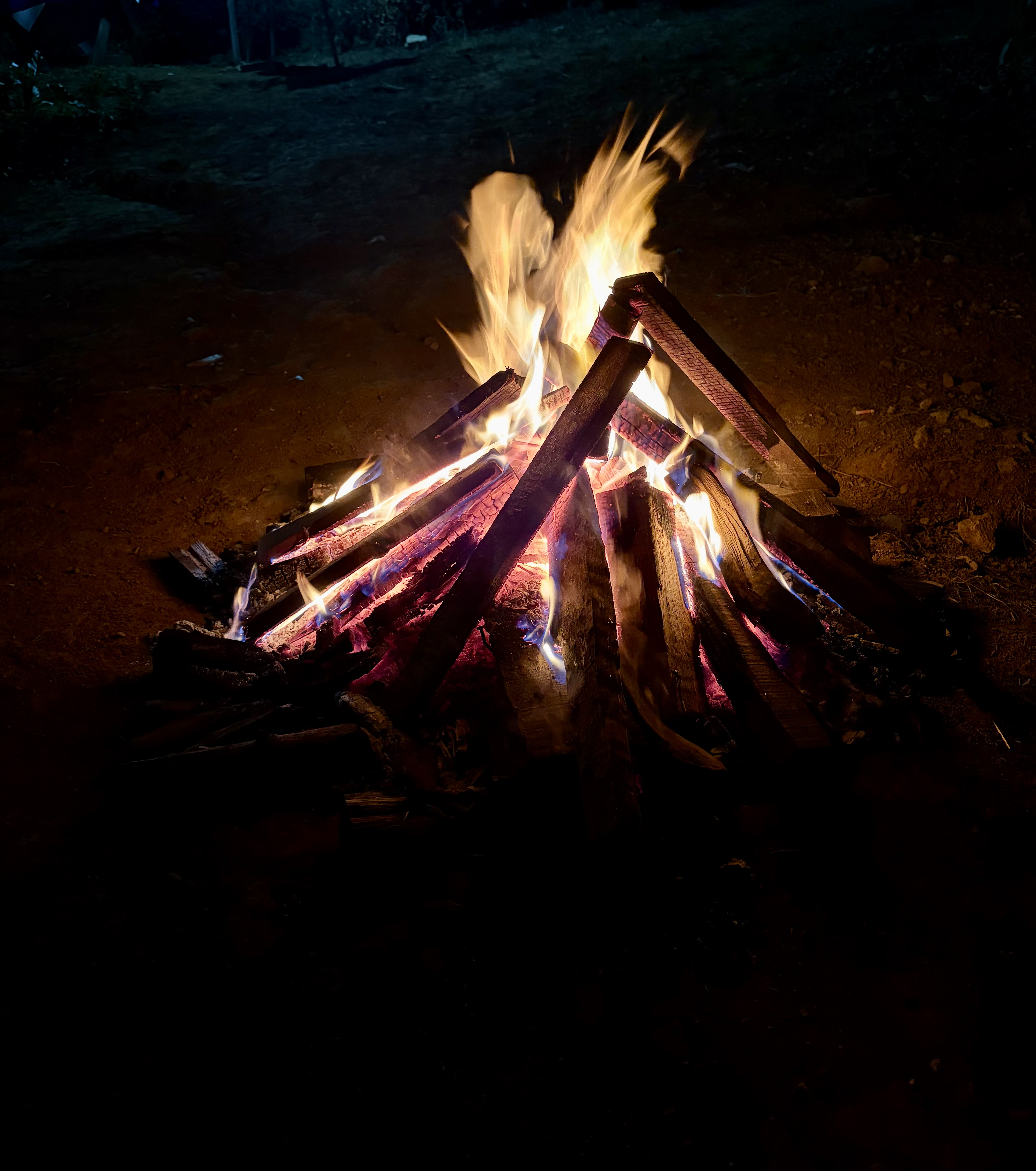 A brightly burning campfire made of stacked wooden logs, with flames flickering upwards against a dark background. The fire emits a warm glow, illuminating the surrounding area slightly, while shadows are cast on the ground.