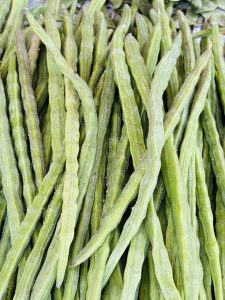 A close-up view of a pile of long, green, cylindrical vegetables, commonly known as drumsticks or moringa pods. The pods are slender, with a ridged texture and a slight sheen, and are arranged overlapping one another, showcasing their elongated shape and vibrant green color.
