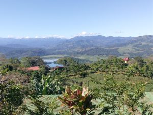 A panoramic view of a lush valley featuring a small lake, red-roofed houses, and rolling green hills under a clear blue sky.