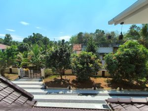 A bright, serene view from above with trees, a patterned driveway, a gated entrance, and buildings partly hidden among greenery under a clear blue sky.