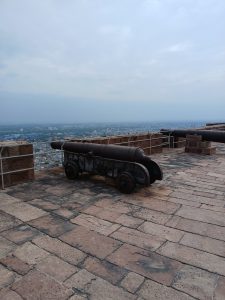Two antique iron cannons on wooden wheeled carriages sitting on a stone fort rampart, overlooking a sprawling city under a hazy sky.