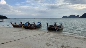 Several wooden longtail boats with colorful decorations are parked on a white sand beach. The ocean water is calm under a cloudy sky. Large green islands are visible in the far distance across the sea.