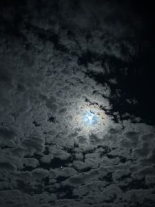 A night sky filled with dark, textured clouds, partially illuminated by a bright, glowing moon. The moon is visible through a break in the clouds, casting a soft glow over the scene. A few faint stars can be seen in the lower part of the image, adding to the tranquil atmosphere.