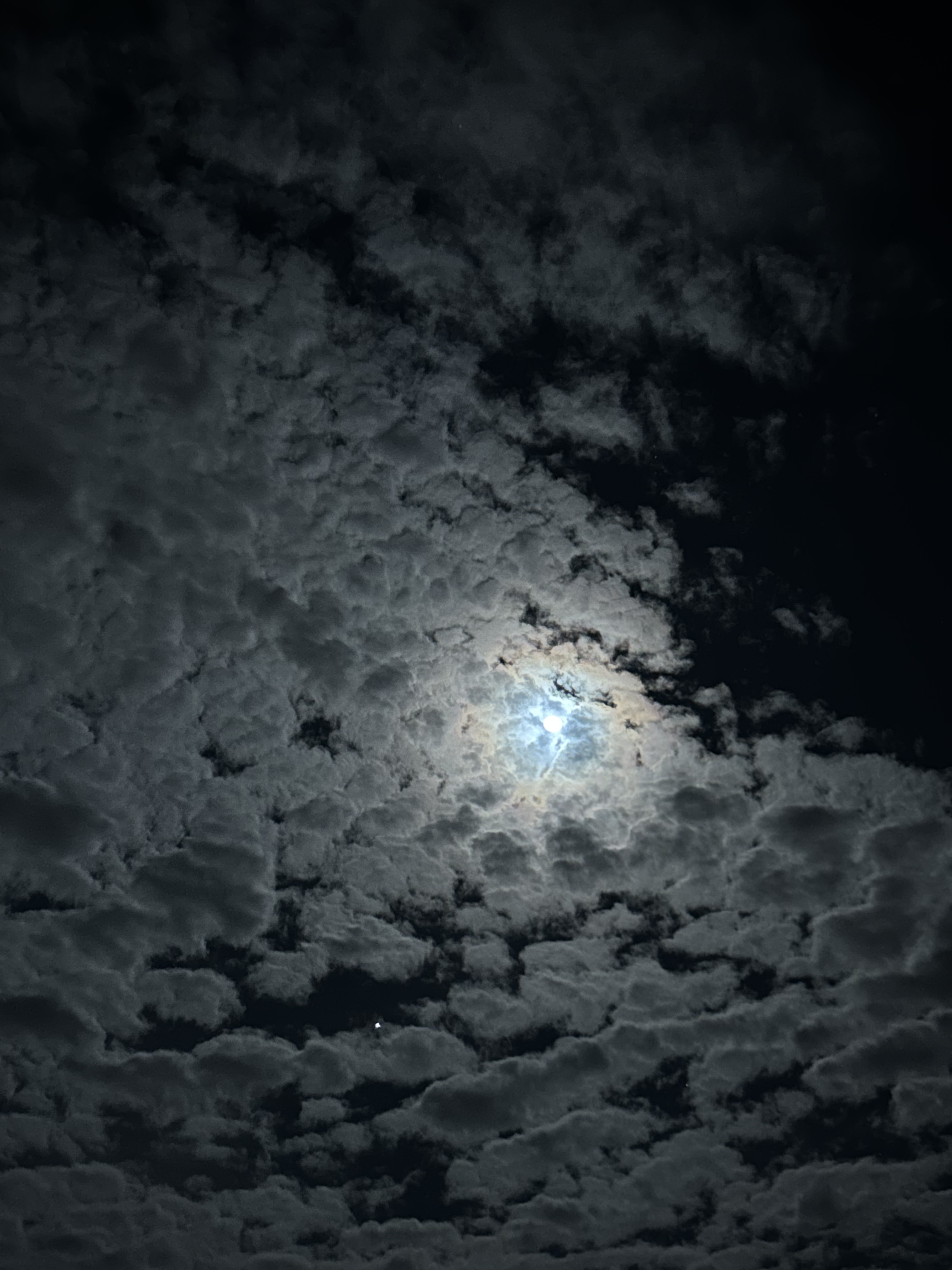A night sky filled with dark, textured clouds, partially illuminated by a bright, glowing moon. The moon is visible through a break in the clouds, casting a soft glow over the scene. A few faint stars can be seen in the lower part of the image, adding to the tranquil atmosphere.