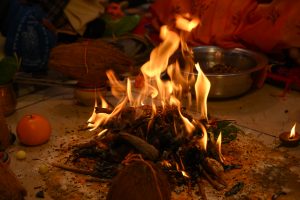 A sacred fire ritual (havan) in progress with flames rising from offerings, surrounded by traditional items in a warm devotional setting.