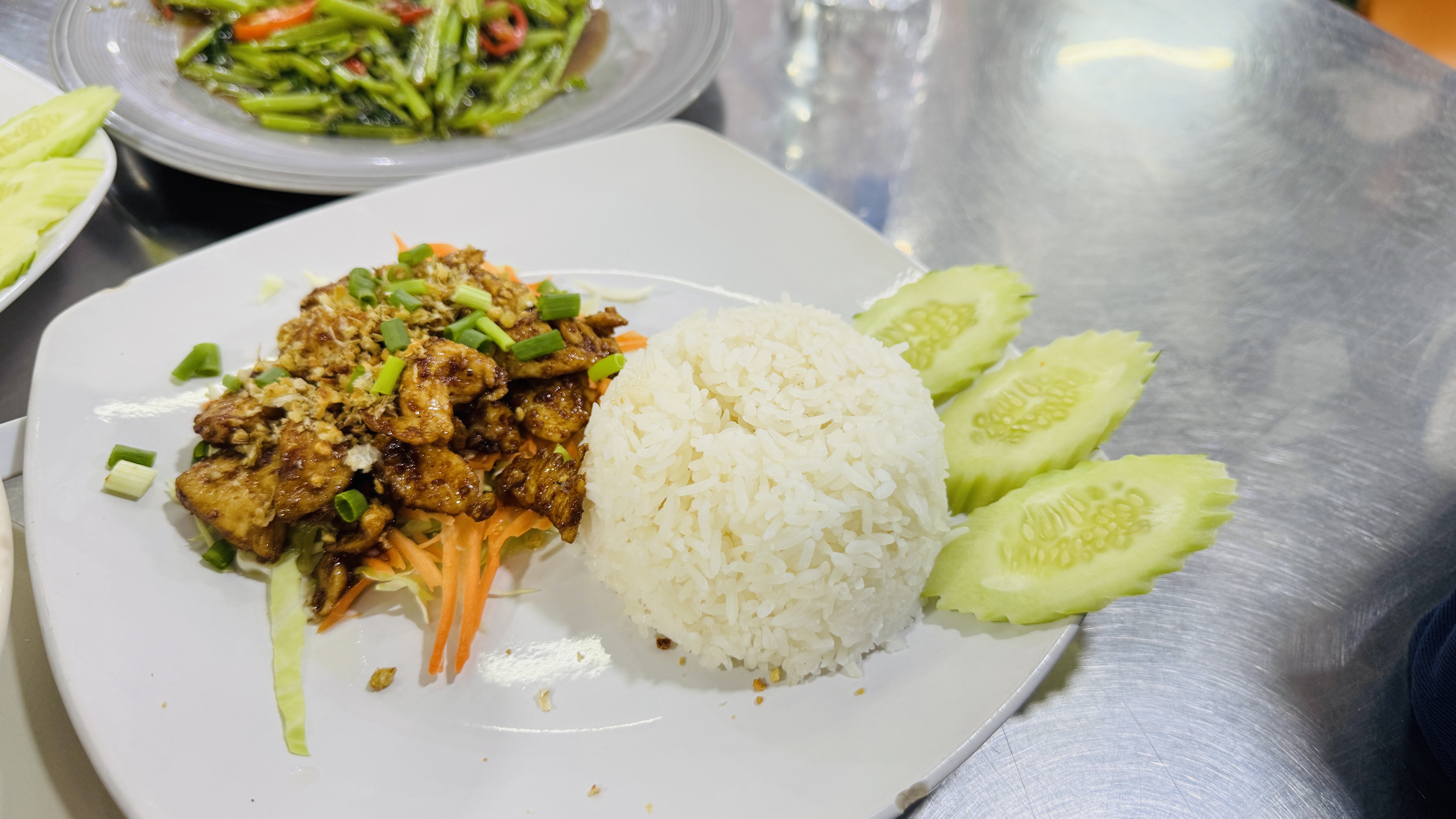 A white plate of stir-fried chicken with garlic and green onions over shredded cabbage and carrots, served with steamed rice and cucumber slices on a shiny metal table in a restaurant.