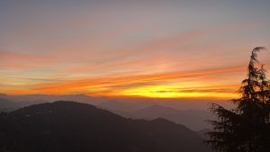 A stunning mountain sunset with an orange-golden sky, misty layered hills, soft clouds on the horizon, and dark trees in the foreground creating a peaceful, cinematic scene.
