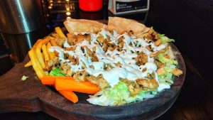 A wooden serving platter holds shawarma, featuring shredded lettuce topped with grilled chicken pieces and drizzled with creamy white sauce.