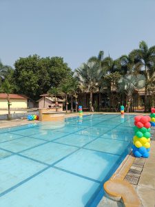 Blue swimming pool with colorful balloon columns along its edge, a fountain, palm trees, and buildings under a clear sky.