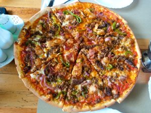 A large, round pizza with a golden-brown, raised crust features melted cheese, savory meat chunks, and a colorful mix of green bell peppers, onions, and cauliflower. The sliced pie sits on a wooden table, with some pieces slightly lifted and various condiments displayed in the background.