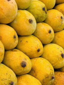 A close-up image of a pile of ripe yellow mangoes, showcasing their smooth skin and small brown spots. The mangoes are arranged closely together, creating a vibrant display of color and texture.
