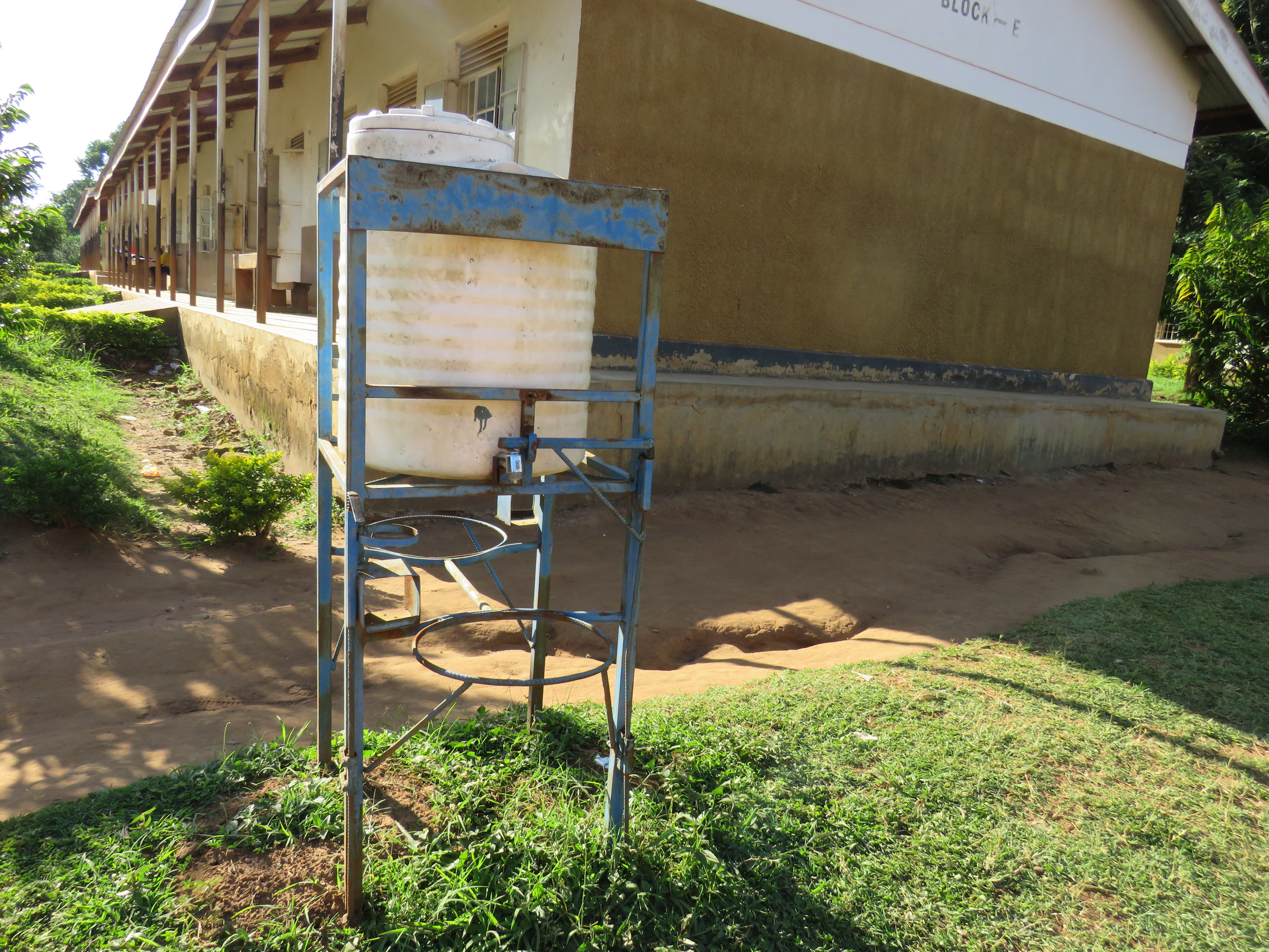 A white water tank on a blue stand sits in grass beside a long building, with a small shrub nearby.