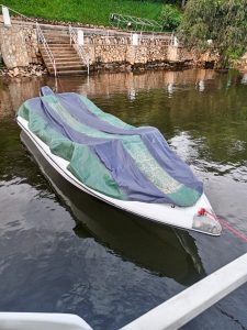 A small white boat covered with a blue and green tarp floating near a stone lakeside wall.
